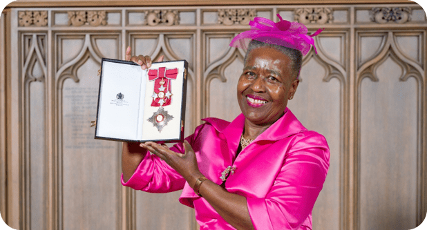 A woman in a bright pink outfit and matching hat smiles while holding up an open box displaying a medal and ribbon. She stands in front of a wooden carved background.