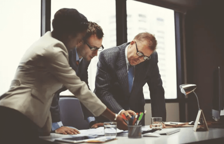 Three business professionals, two men and one woman, are gathered around a desk reviewing documents in a well-lit office with large windows.