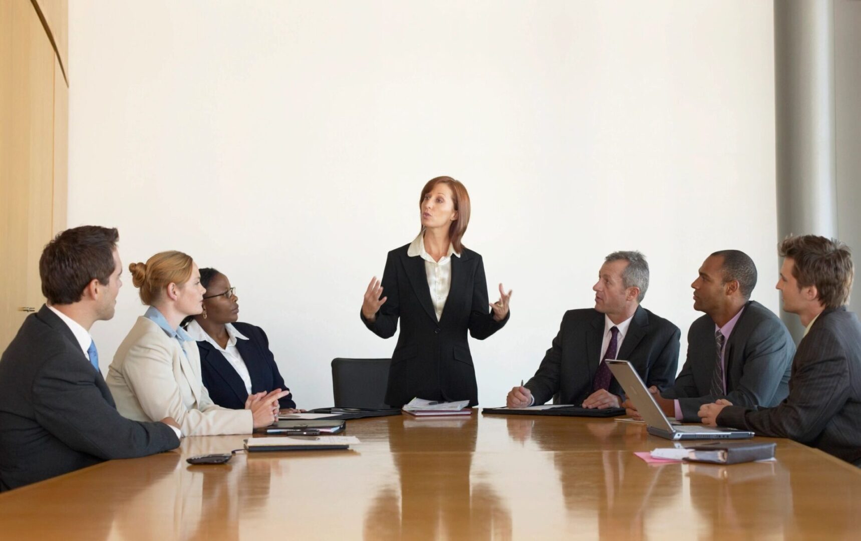 A woman in a suit stands and speaks to six seated colleagues during a business meeting in a conference room.