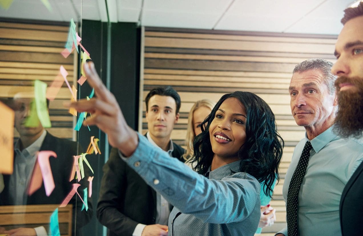 Several people in business attire collaborate in an office, with one woman pointing at sticky notes on a glass wall.