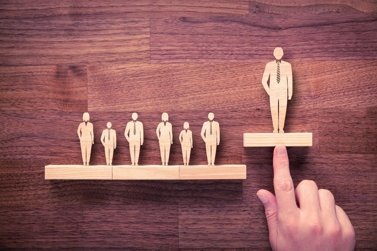 A hand supports a wooden block elevating a wooden figure wearing a tie, while five similar figures stand on a horizontal block on a wooden surface.