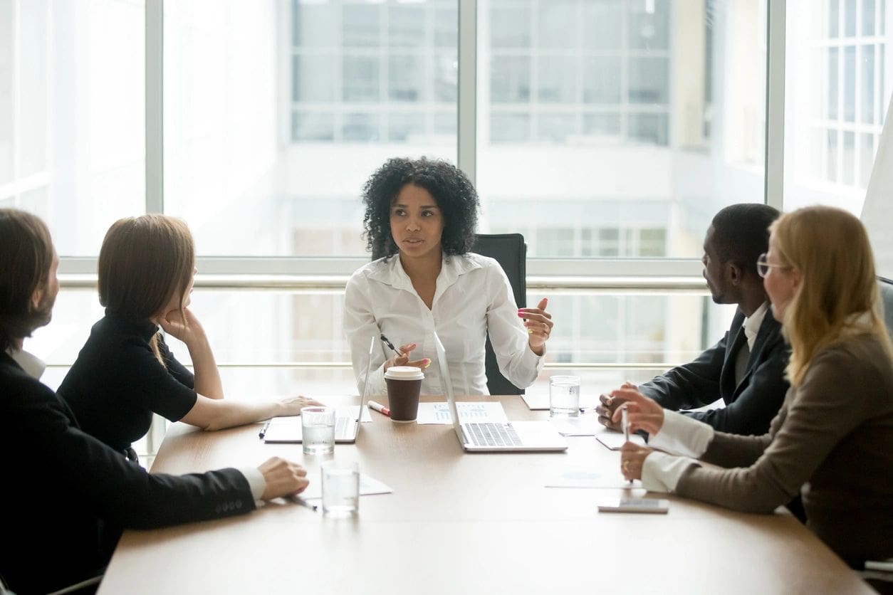 A woman leads a meeting at a conference room table, speaking to four seated colleagues. The table has laptops, documents, and drinks with large windows in the background.