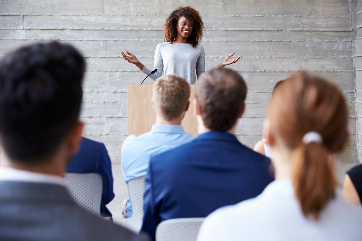 A woman stands at a podium, speaking to a seated audience in a conference setting.